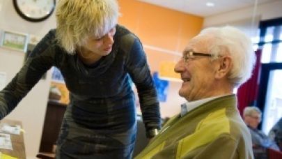 Zorgmedewerkster in gesprek met een oudere heer in een zaal waar meedere ouderen aanwezig zijn