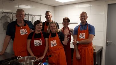Groepsfoto van Facilicom-medewerkers die in de keuken staan bij Resto van Harte. Allen met een rood Resto van Harte schort.