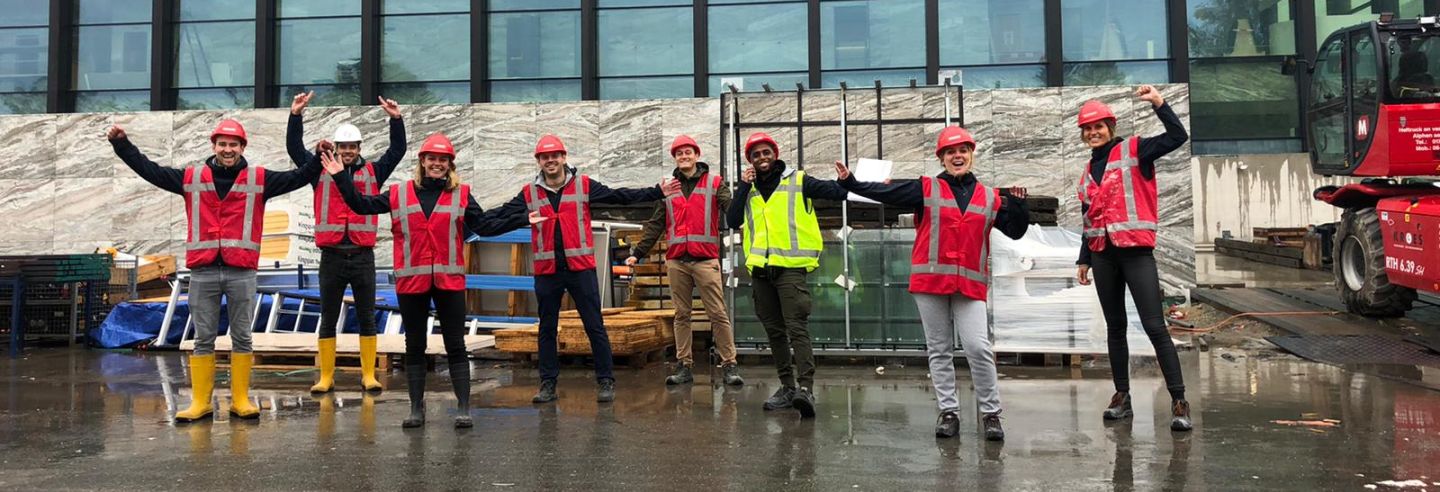 Een groepsfoto van de Facilicom Group trainees op een bouwplaats oranje hesjes aan en een bouwhelm op.