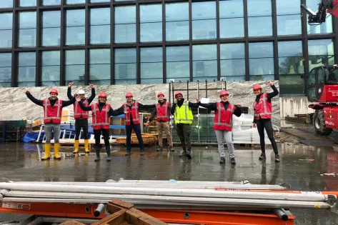 Een groepsfoto van de Facilicom Group trainees op een bouwplaats oranje hesjes aan en een bouwhelm op.
