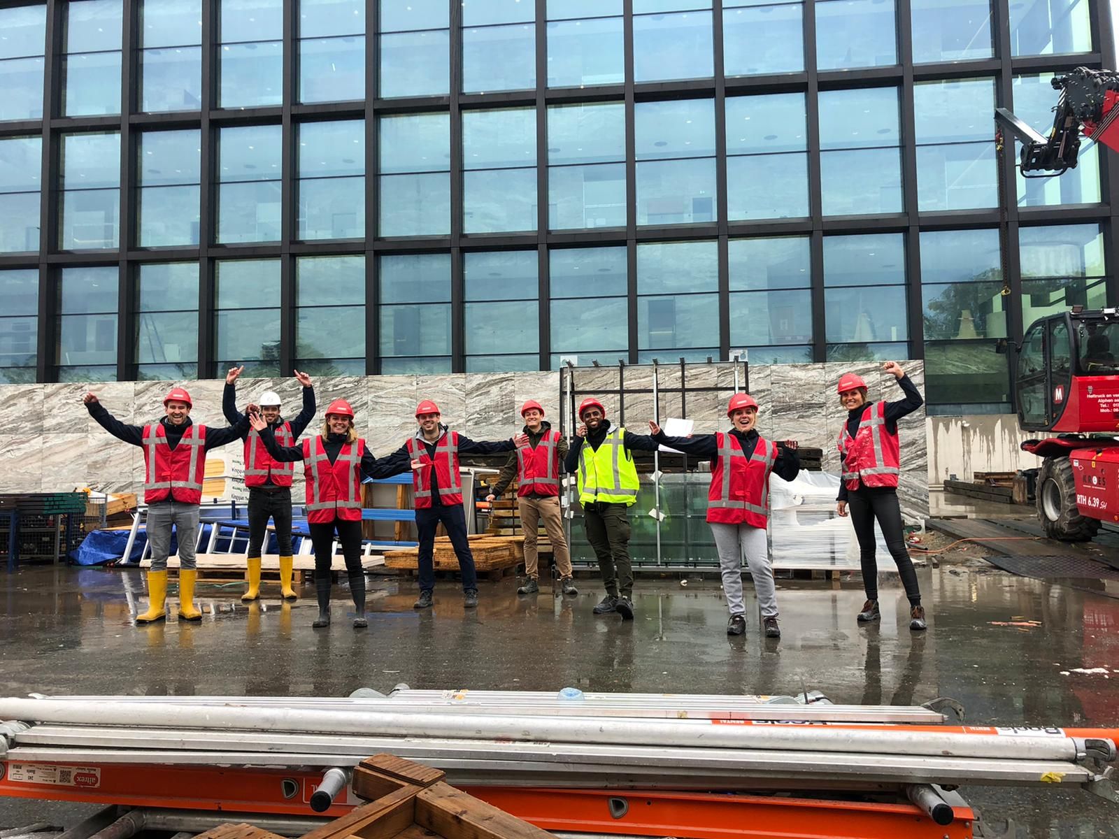 Een groepsfoto van de Facilicom Group trainees op een bouwplaats oranje hesjes aan en een bouwhelm op.